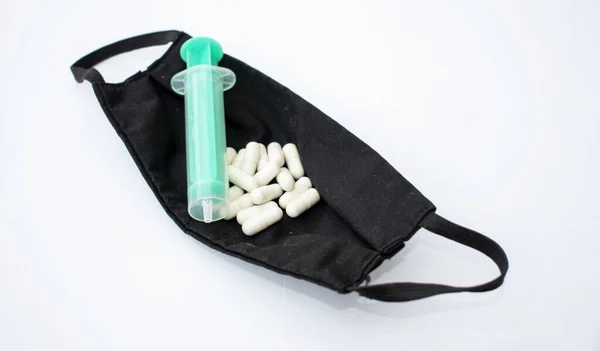 Bir sürü hap ve yeşil şırınga beyaz bir arka planda siyah, kumaş bir yüz maskesi içinde