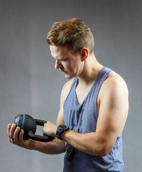 Egzersiz yaparken siyah bir çaydanlığa bakan beyaz bir adam. Suratı asık, odaklanmış. Demiri iki elinde tutuyor.,