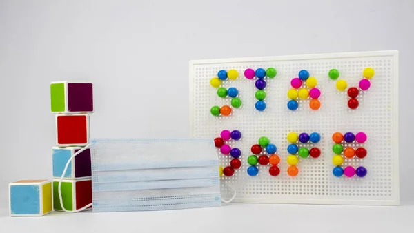 Renkli bloklar, Güvenli Kal tabelasına yerleştirilmiş iğneler ve beyaz bir arka planda cerrahi maske..