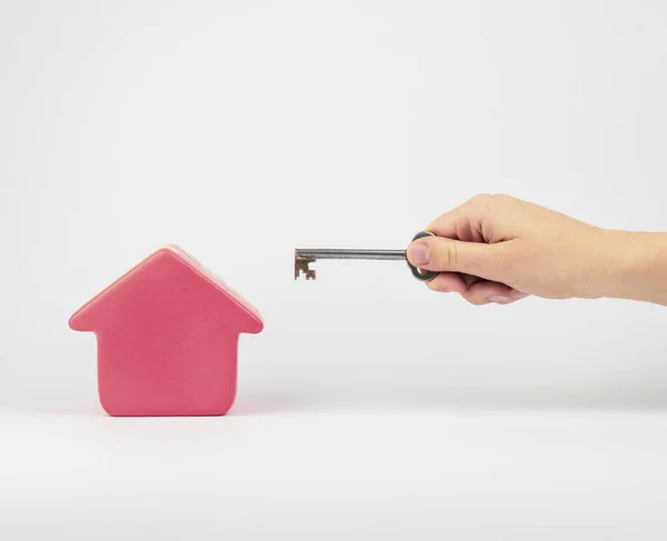 Gümüş renkli, şık bir anahtarı tutan ve küçük kırmızı bir ev heykelciğine doğrultan bir kadın eli. Resmin beyaz bir arka planı var ve bir ev satın alma, sunma veya kiralama metaforunu temsil ediyor.