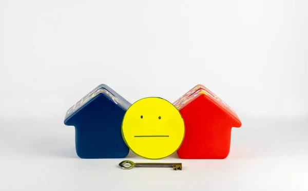 İki ev, anahtar ve sarı bir yüz belirsiz bir emlak piyasasını temsil ediyor. Tarafsız yüz, potansiyel alıcıları piyasada bir krizin eşiğinde oldukları için ilk evlerini satın alma konusunda uyardı