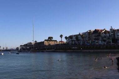 Portekiz 'in Cascais kentindeki pitoresk kıyı şeridinin güneşli geniş açılı bir görüntüsü körfezin sakin sularına odaklanıyor, ayrıca Fishermens Beach Praia dos Pescadores veya Cascais Körfezi olarak da bilinir. Ön planda, insanların yüzdüğü derin mavi deniz yer alıyor.