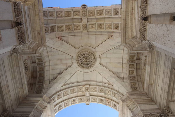 A symmetrical low-angle perspective of the ornate limestone interior of the Rua Augusta Arch. The image showcases the architectural beauty of the Pombaline style, featuring decorative floral rosettes, rectangular stone coffers, and a central circular