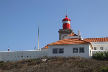 Portekiz 'in Cabo da Roca kentindeki turuncu terakotta kiremit çatılı beyaz duvarlı binaların üzerinde parlak kırmızı bir deniz feneri kulesi bulunuyor. Sahne güneşli bir günde, açık mavi bir gökyüzü ile Avrupa 'nın en batı noktasını vurguluyor.