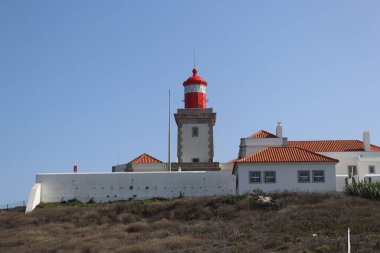 Portekiz 'in Cabo da Roca kentindeki turuncu terakotta kiremit çatılı beyaz duvarlı binaların üzerinde parlak kırmızı bir deniz feneri kulesi bulunuyor. Sahne güneşli bir günde, açık mavi bir gökyüzü ile Avrupa 'nın en batı noktasını vurguluyor.