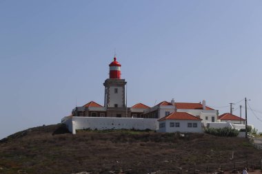 Portekiz 'in Cabo da Roca kentindeki turuncu terakotta kiremit çatılı beyaz duvarlı binaların üzerinde parlak kırmızı bir deniz feneri kulesi bulunuyor. Sahne güneşli bir günde, açık mavi bir gökyüzü ile Avrupa 'nın en batı noktasını vurguluyor.