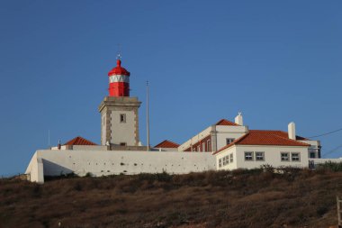 Portekiz 'in Cabo da Roca kentindeki turuncu terakotta kiremit çatılı beyaz duvarlı binaların üzerinde parlak kırmızı bir deniz feneri kulesi bulunuyor. Sahne güneşli bir günde, açık mavi bir gökyüzü ile Avrupa 'nın en batı noktasını vurguluyor.
