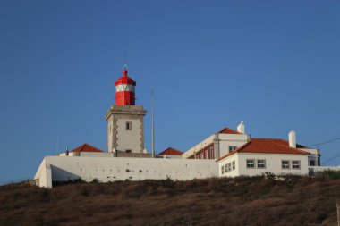 Portekiz 'in Cabo da Roca kentindeki turuncu terakotta kiremit çatılı beyaz duvarlı binaların üzerinde parlak kırmızı bir deniz feneri kulesi bulunuyor. Sahne güneşli bir günde, açık mavi bir gökyüzü ile Avrupa 'nın en batı noktasını vurguluyor.
