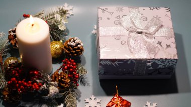 Büyük yanan beyaz bir mum, çam dalları ve Noel oyuncakları hediyeleri, toplar, Rowan, çam kozalağı ve arka planda kar taneleri olan büyük gümüş bir hediye kutusu.       