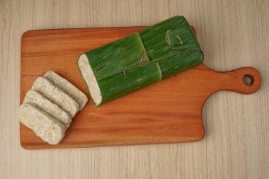 Tempeh muz yapraklarına sarılmış ve ahşap kesme tahtasının üzerinde tempeh dilimleri var.
