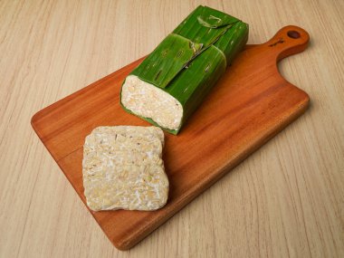 Tempeh muz yapraklarına sarılmış ve ahşap kesme tahtasının üzerinde tempeh dilimleri var.