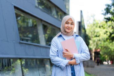 Başörtüsü takan bir kadın pembe bir kitap tutuyor. Gülümsüyor ve mutlu.