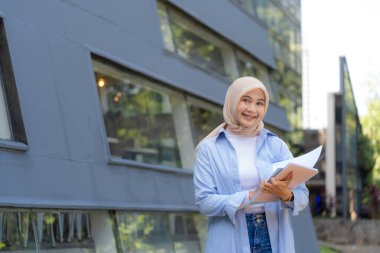 Mavi gömlekli ve beyaz eşarplı bir kadın kitap okuyor. Bir üniversitenin önünde duruyor.