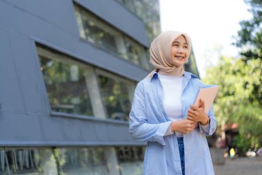 Başörtüsü takan bir kadın pembe bir kitap tutuyor. Gülümsüyor ve mutlu.