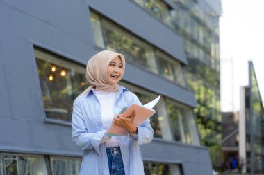 Mavi gömlekli ve beyaz eşarplı bir kadın kitap okuyor. Bir üniversitenin önünde duruyor.