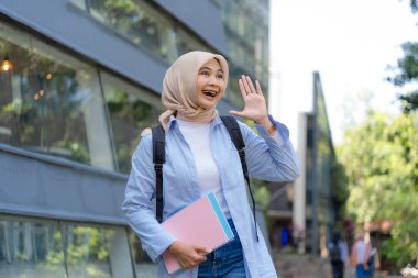 Mavi gömlek ve başörtüsü takan genç bir kadın pembe bir kitap ve sırt çantası tutarken birine el sallıyor.