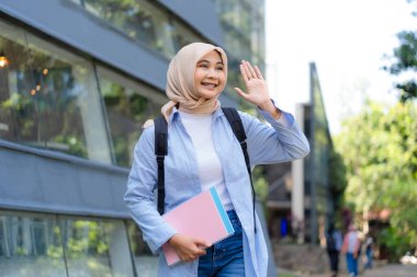 Mavi gömlek ve başörtüsü takan genç bir kadın pembe bir kitap ve sırt çantası tutarken birine el sallıyor.