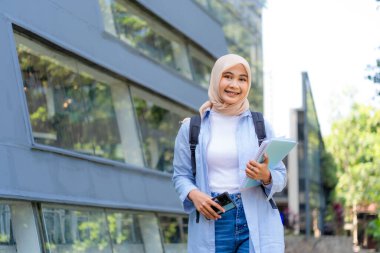 Mavi gömlek ve beyaz gömlek giyen genç bir kadın bir cep telefonu ve bir kitap tutuyor. Gülümsüyor ve mutlu.