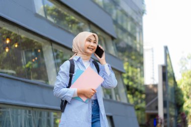 Bir yığın kitap tutarken tesettürlü genç bir kadın cep telefonuyla konuşuyor. Keyfi yerinde ve sohbetten zevk alıyor.