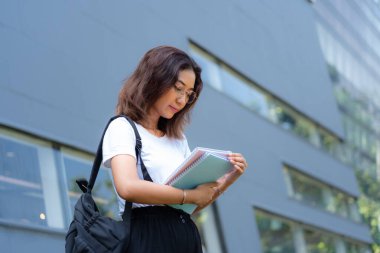 Gözlüklü ve beyaz gömlekli bir kadın bir kitap ve bir sırt çantası tutuyor. Büyük bir penceresi olan bir binanın önünde duruyor.