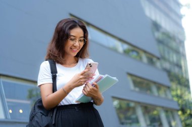 Bir kadın elinde not defteri tutarken telefonuna bakıyor. O, gözlük ve beyaz bir gömlek giyiyor.