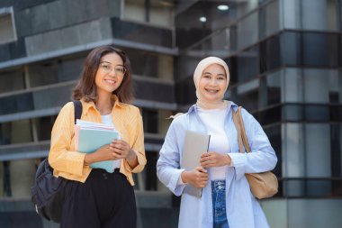 İki genç kadın gülümsüyor ve ellerinde kitap ve tabletlerle. Sırt çantası takıyorlar ve öğrenci gibi görünüyorlar.