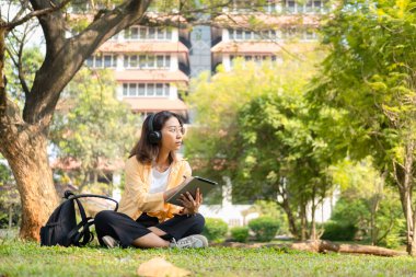 Modern bir Asyalı öğrenci, online eğitim ve yaratıcılığı simgeleyen üniversite parkındaki bir ağacın altında otururken not almak için dijital tablet kullanır..