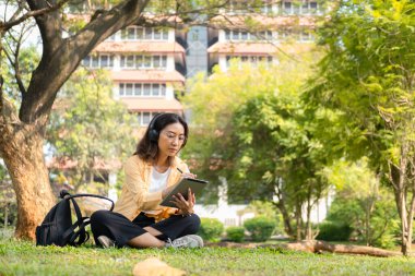 Modern bir Asyalı öğrenci, online eğitim ve yaratıcılığı simgeleyen üniversite parkındaki bir ağacın altında otururken not almak için dijital tablet kullanır..