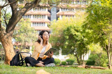 Modern bir Asyalı öğrenci, online eğitim ve yaratıcılığı simgeleyen üniversite parkındaki bir ağacın altında otururken not almak için dijital tablet kullanır..