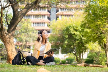 Modern bir Asyalı öğrenci, online eğitim ve yaratıcılığı simgeleyen üniversite parkındaki bir ağacın altında otururken not almak için dijital tablet kullanır..