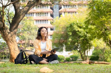 Modern bir Asyalı öğrenci, online eğitim ve yaratıcılığı simgeleyen üniversite parkındaki bir ağacın altında otururken not almak için dijital tablet kullanır..