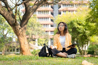 Modern bir Asyalı öğrenci, online eğitim ve yaratıcılığı simgeleyen üniversite parkındaki bir ağacın altında otururken not almak için dijital tablet kullanır..