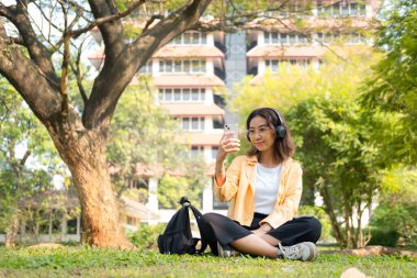 Kablosuz kulaklık takan genç bir kız öğrenci çimlerin üzerinde tablet ve sırt çantasıyla oturup ağacın altında sakin bir çalışma saatinin keyfini çıkarıyor..