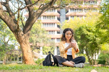 Kablosuz kulaklık takan genç bir kız öğrenci çimlerin üzerinde tablet ve sırt çantasıyla oturup ağacın altında sakin bir çalışma saatinin keyfini çıkarıyor..