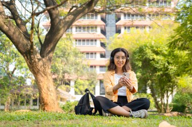 Kablosuz kulaklık takan genç bir kız öğrenci çimlerin üzerinde tablet ve sırt çantasıyla oturup ağacın altında sakin bir çalışma saatinin keyfini çıkarıyor..