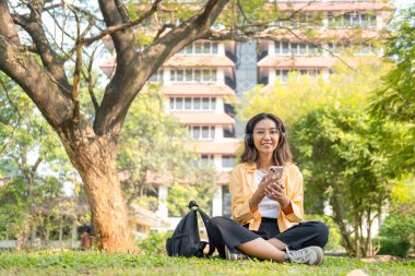 Kablosuz kulaklık takan genç bir kız öğrenci çimlerin üzerinde tablet ve sırt çantasıyla oturup ağacın altında sakin bir çalışma saatinin keyfini çıkarıyor..