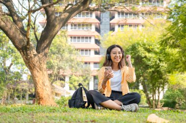 Kablosuz kulaklık takan genç bir kız öğrenci çimlerin üzerinde tablet ve sırt çantasıyla oturup ağacın altında sakin bir çalışma saatinin keyfini çıkarıyor..