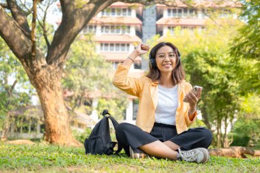 Kablosuz kulaklık takan genç bir kız öğrenci çimlerin üzerinde tablet ve sırt çantasıyla oturup ağacın altında sakin bir çalışma saatinin keyfini çıkarıyor..