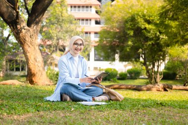 Başörtüsü takan genç bir Müslüman kadın açık havada tablet ve açık defterle çalışıyor. Ağaçlarla çevrili ve arka planda bir üniversite binası var..