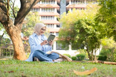 Başörtüsü takan genç bir Müslüman kadın açık havada tablet ve açık defterle çalışıyor. Ağaçlarla çevrili ve arka planda bir üniversite binası var..