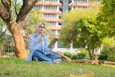 Başörtüsü takan düşünceli Müslüman öğrenci, eğitimde yaratıcılığı ve problem çözmeyi temsil eden not defterleriyle çimlerin üzerinde otururken bir tablet ve kalem tutuyor..