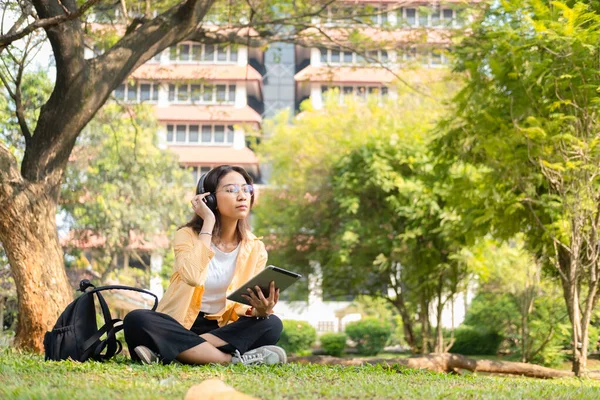 Modern bir Asyalı öğrenci, online eğitim ve yaratıcılığı simgeleyen üniversite parkındaki bir ağacın altında otururken not almak için dijital tablet kullanır..