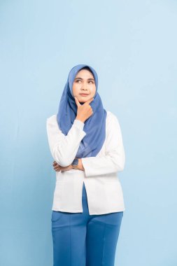 Düşünceli genç bir Asyalı Müslüman kadının portresi, çenesi elinde, yukarı bakıyor ve mavi bir arka plana bakıyor..