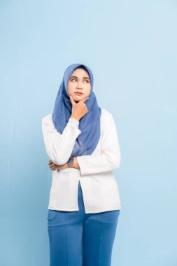 Düşünceli genç bir Asyalı Müslüman kadının portresi, çenesi elinde, yukarı bakıyor ve mavi bir arka plana bakıyor..