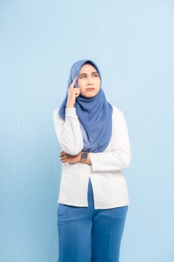 Düşünceli genç bir Asyalı Müslüman kadının portresi, çenesi elinde, yukarı bakıyor ve mavi bir arka plana bakıyor..
