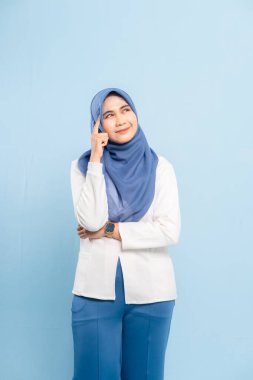 Düşünceli genç bir Asyalı Müslüman kadının portresi, çenesi elinde, yukarı bakıyor ve mavi bir arka plana bakıyor..