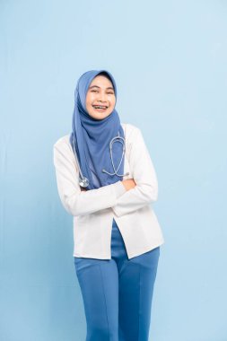 Kendinden emin ve gülümseyen genç bir Asyalı Müslüman doktorun ya da steteskoplu bir hemşirenin portresi, kolları mavi bir arka planda izole edilmiş..