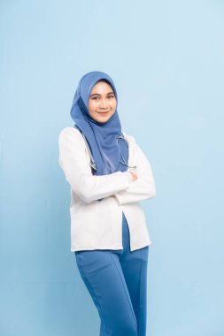 Kendinden emin ve gülümseyen genç bir Asyalı Müslüman doktorun ya da steteskoplu bir hemşirenin portresi, kolları mavi bir arka planda izole edilmiş..