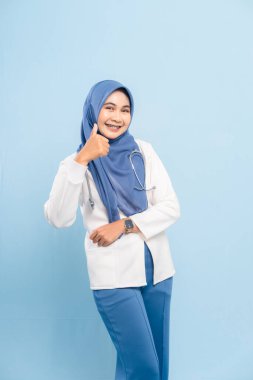 Gülümseyen genç bir Asyalı Müslüman doktor ya da hemşirenin portresi, mavi bir arka planda izole edilmiş, onay ya da iyi sağlığı ifade eden,.