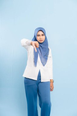 Ciddi bir Asyalı genç Müslüman doktor ya da hemşirenin portresi, mavi arka planda izole edilmiş, onaylanmamış ya da olumsuz teşhisi işaret eden bir başparmak işareti veriyor..